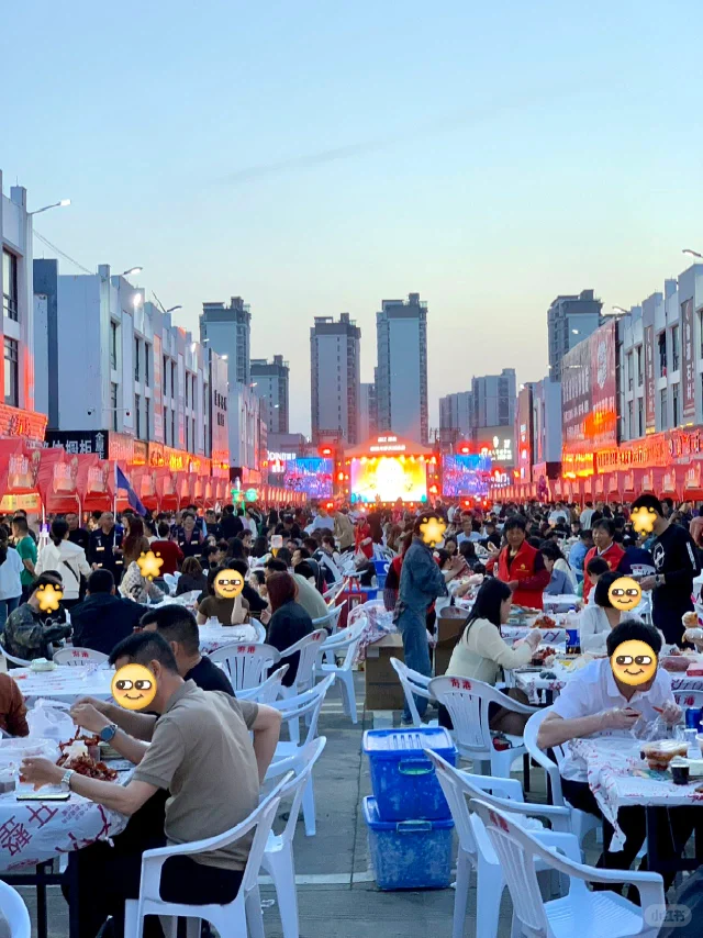 夜晚的定海街头夜市,一张木桌上摆放着一大盆红亮油润的小龙虾,旁边配有冰镇啤酒、一次性手套和纸巾,食客正在开心地剥虾进食,背景有灯光和人群 定海夜市小龙虾大餐全景