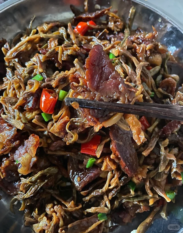 一位家庭主妇在明亮的现代厨房中,正在用铁锅翻炒已泡发并焯水后的豆角丝,锅中搭配有腊肉片、蒜末和青红椒丝,香气四溢。灶台旁摆放着泡发前的干豆角丝样本和温水浸泡容器,形成鲜明对比,体现从干品到美食的转变过程 家庭厨房中正在烹饪泡发好的豆角丝菜肴