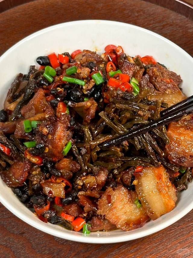 一名厨师正在灶台前将泡发好的干豆角切段,倒入热油锅中与五花肉一同翻炒,锅中升腾起浓郁香气,旁边放着泡发前后的对比样品,展示干豆角的吸水膨胀特性 厨房中用干豆角烹饪菜肴的过程