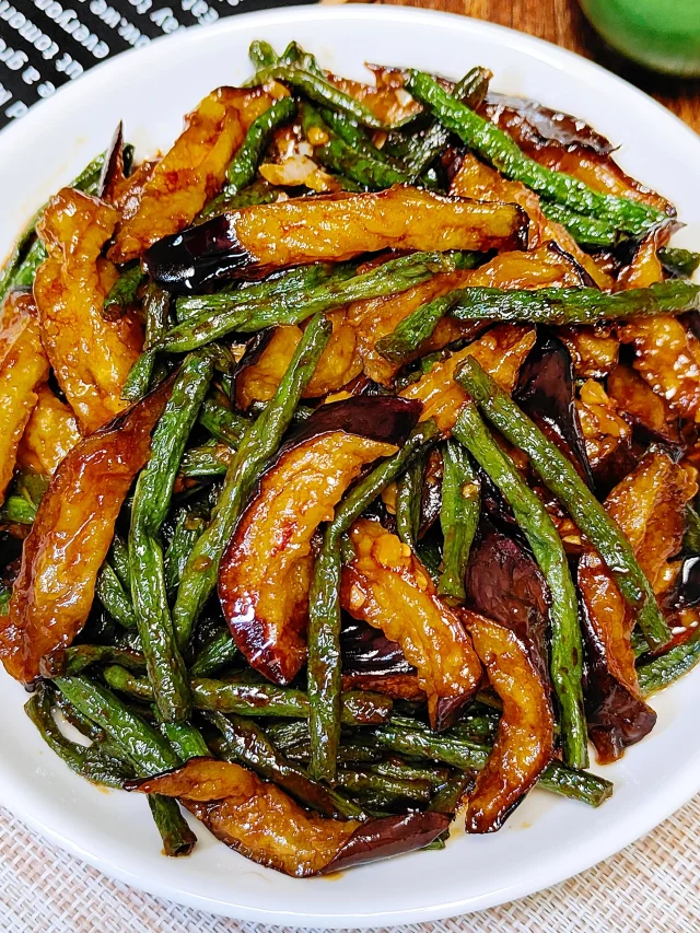 一口黑色铸铁炒锅中正在翻炒着油亮的豆角与茄子，食材色泽分明，豇豆翠绿，茄子深紫泛光，蒸汽升腾