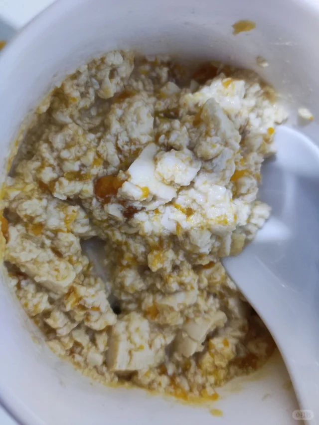 白色瓷碗中盛放着顺滑浓稠的金黄色南瓜豆浆羹,表面泛着淡淡油光,旁边摆放木勺和几粒黑色亚麻籽点缀,背景虚化的厨房环境中可见蒸锅与豆浆机 南瓜豆浆羹特写