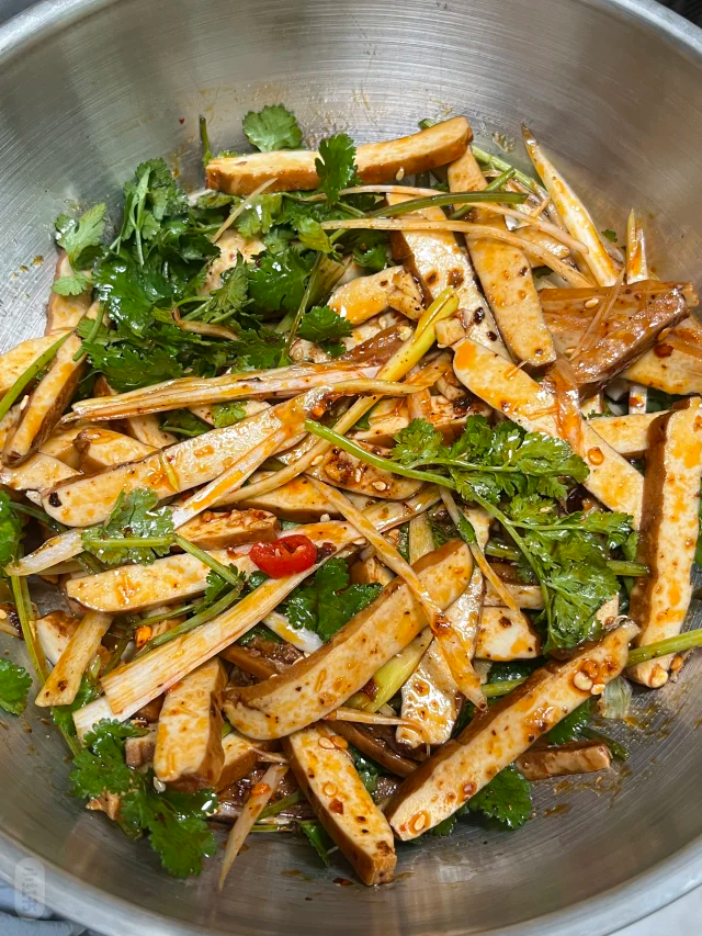 厨房操作台上的香菜拌豆干制作过程,左侧为切好的大叶香菜段和豆干条,中间为加了调料的混合食材,右侧为正在倒入热芝麻油的瞬间,使用小铜勺倾倒,背景为木质砧板和调味瓶 香菜拌豆干制作过程图