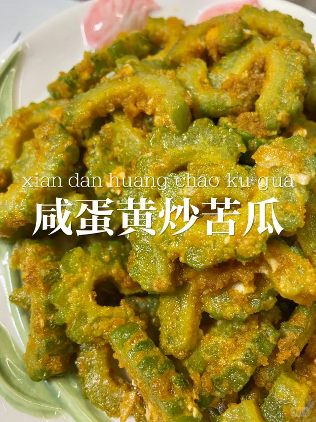 新鲜翠绿的长条苦瓜一根置于木砧板左侧,右侧摆放两个完整的灰白色咸鸭蛋,背景有剥开的咸鸭蛋特写,蛋黄呈橙红色油润状态,旁边有三瓣去皮大蒜 苦瓜与咸鸭蛋食材摆盘