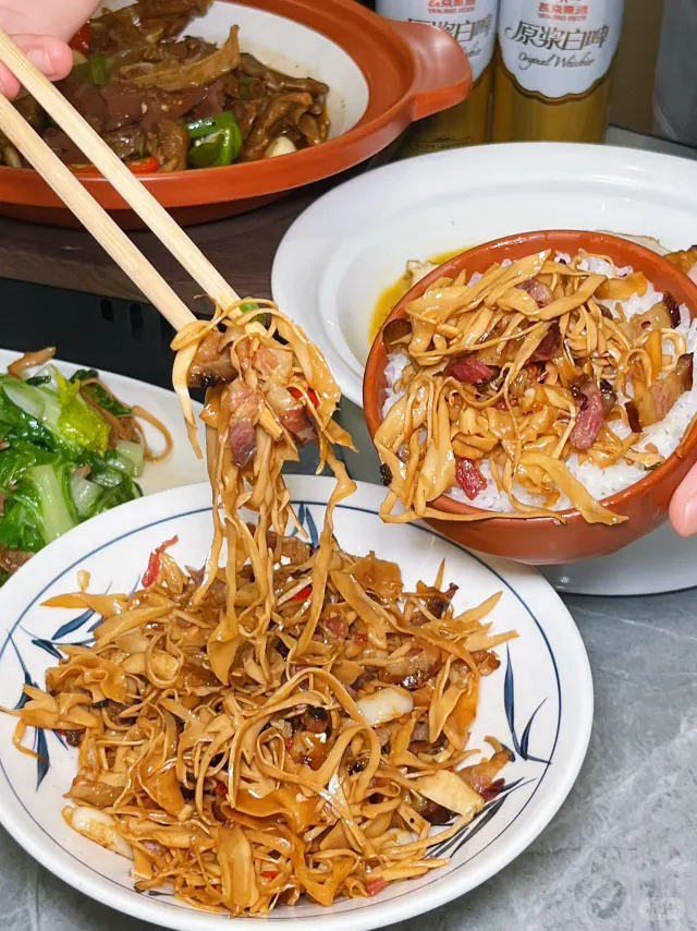 一盘热气腾腾的竹笋炒腊肉被端上木制餐桌，菜肴色泽金黄油亮，腊肉与笋片交织，旁边配有米饭和小碟酱菜