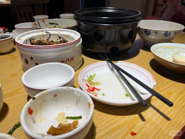 一个现代厨房场景,女性正在将蒸好的黄心红薯盛入陶瓷碗中,旁边放有一瓶未开封的白酒,用红色箭头标示‘分开食用’,墙上挂有时钟显示时间为晚上6点30分,强调用餐时间管理的重要性 图片描述
